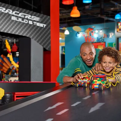 A dad and his son are building a LEGO race car together at LEGOLAND Discovery Centre Toronto. The dad is helping his son assemble the car using colorful LEGO bricks, while both focus on the creative process. The scene is set within a bright, interactive area of the center, with other LEGO structures and activities visible in the background. The father and son share a moment of excitement and teamwork as they work on their race car creation.